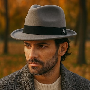 Könnte beinhalten: Grauer Fedora-Hut mit schwarzem Band. Der Hut wird mit einem grauen Fischgrätenmantel und einem cremefarbenen Pullover getragen. Der Hintergrund zeigt verschwommenes Herbstlaub.