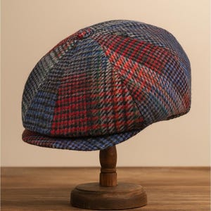 May include: A tweed newsboy cap in a patchwork design. The cap features a mix of red, blue, and grey plaid patterns. It is displayed on a small wooden stand. The brim is a solid blue. The cap is a classic style.