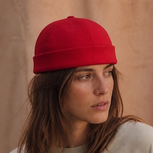 May include: A woman wearing a red beanie hat and a light green t-shirt. The hat is a solid, vibrant red colour. The t-shirt is a neutral, muted green. The woman has long, light brown hair.
