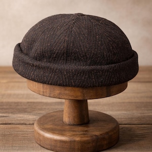 Puede incluir: Un gorro de color marrón oscuro y texturizado con un borde enrollado, exhibido en un soporte de madera. El gorro tiene una corona redondeada y un sutil patrón de espiga. El soporte es de madera con un diseño clásico.
