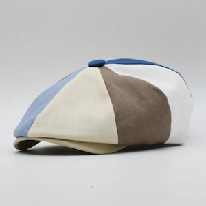 May include: A patchwork newsboy cap in a variety of colors, including light blue, cream, brown, white, and navy blue. The cap has a small, round button on top and a curved brim. The hat is made of a textured fabric.