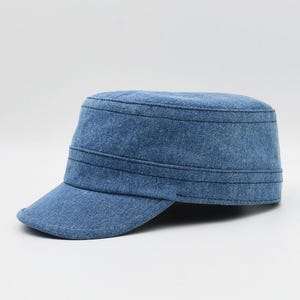 May include: A blue denim cadet cap with a short brim. The cap has a flat top and a structured design, featuring horizontal stitched lines around the crown. The fabric appears to be a light-wash denim, offering a casual and versatile style.