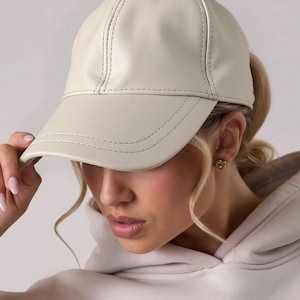 May include: Cream-colored baseball cap with a curved brim. The cap is made of a smooth material and features stitched detailing along the seams and brim. A classic, stylish accessory.