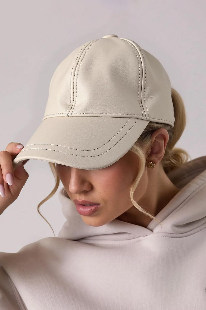 May include: Cream-colored baseball cap with a curved brim. The cap is made of a smooth material and features stitched detailing along the seams and brim. A classic, stylish accessory.