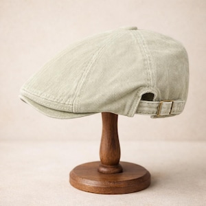 Puede incluir: Gorra de estilo newsboy de pana verde claro con una visera corta. La gorra tiene una correa de tela con una hebilla metálica para ajustarla. El sombrero se exhibe en un soporte de madera.