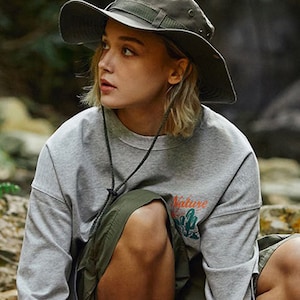 May include: A woman wearing a grey t-shirt with the text "Nature" printed on it, a green skirt, and a green wide-brimmed hat.