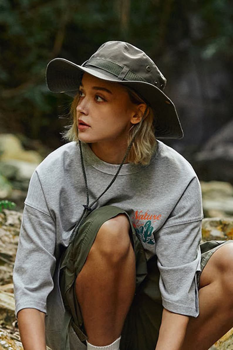 May include: A woman wearing a grey t-shirt with the text "Nature" printed on it, a green skirt, and a green wide-brimmed hat.