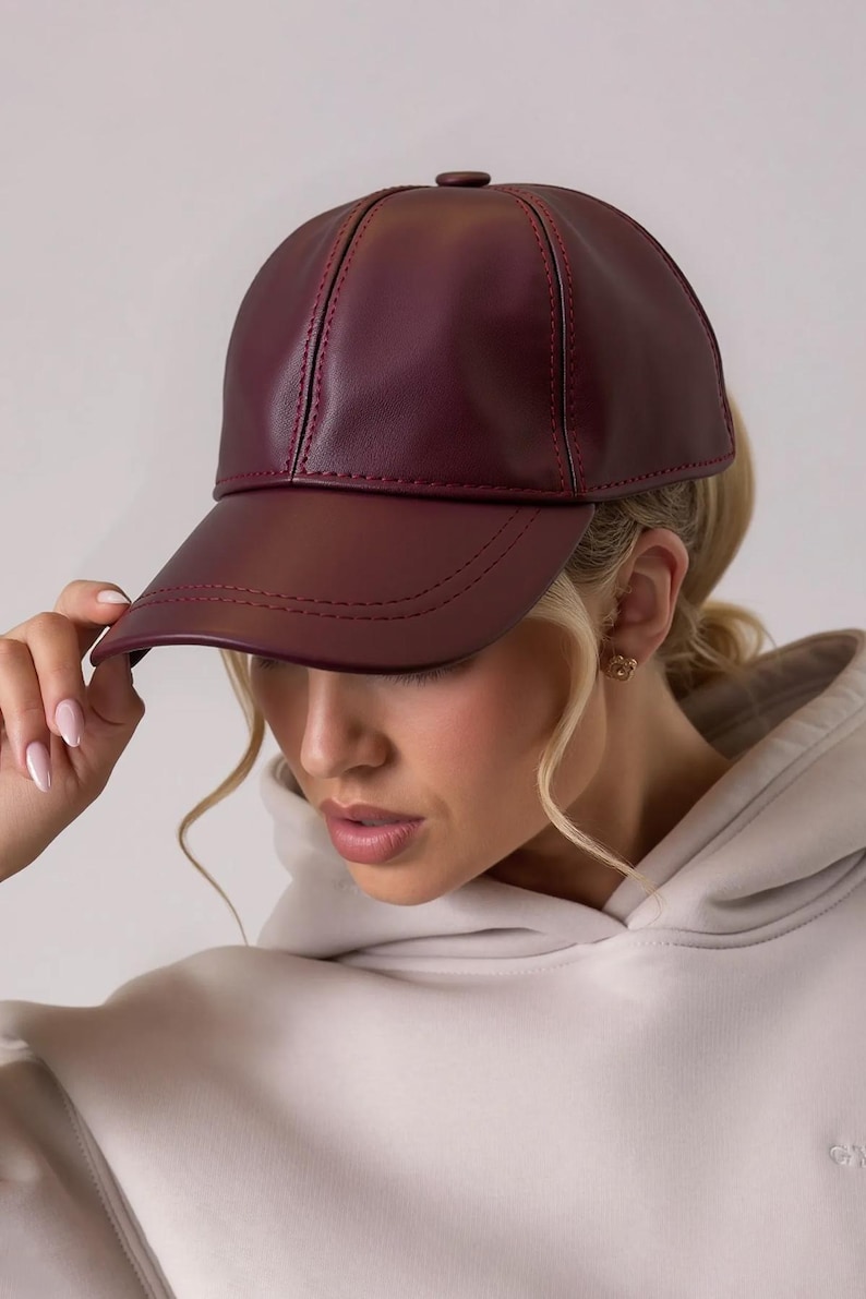 May include: A burgundy baseball cap made of a smooth material. The cap has a curved brim and a structured crown with visible stitching. The cap is being held by a hand with manicured nails.