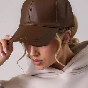 May include: A brown leather baseball cap with a curved brim. The cap has a classic six-panel design with a button on top and stitched detailing. The cap is a solid color and appears to be made of a smooth material.