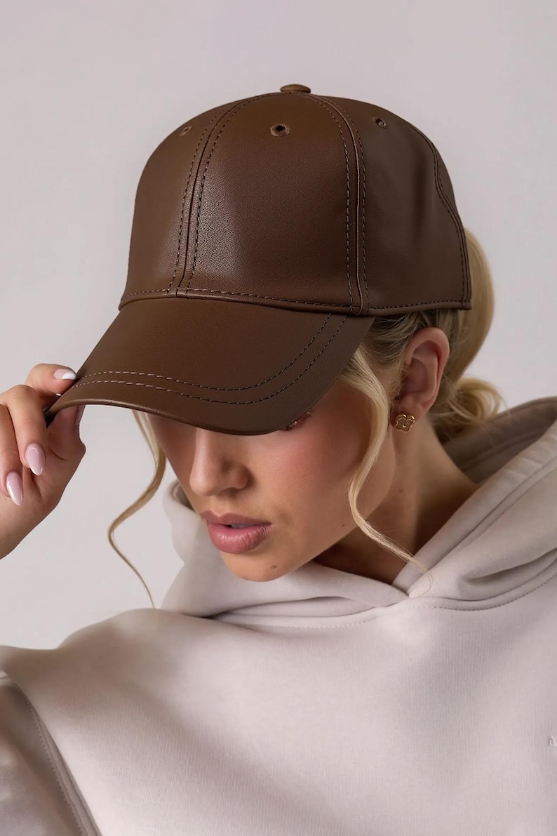 May include: A brown leather baseball cap with a curved brim. The cap has a classic six-panel design with a button on top and stitched detailing. The cap is a solid color and appears to be made of a smooth material.