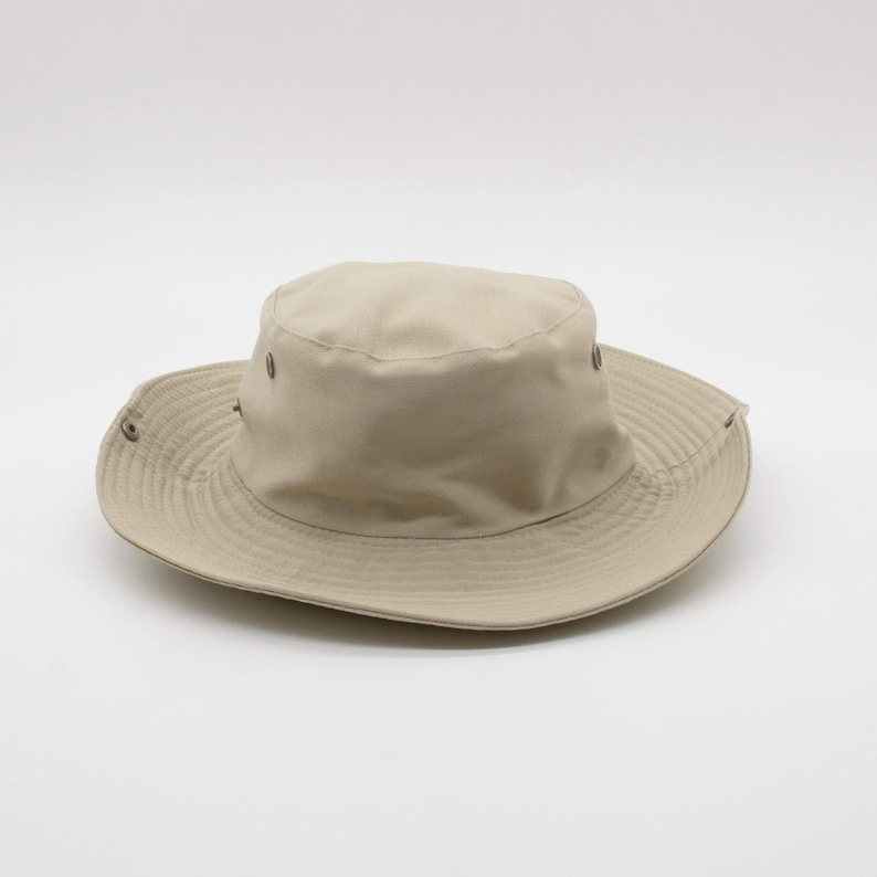 May include: A beige sun hat with a wide brim and a rounded crown. The hat features a series of small metal eyelets around the crown and a structured brim. The hat is designed to provide shade and protection from the sun.