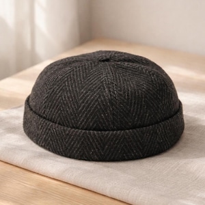 Può includere: Un berretto grigio scuro con motivo a spina di pesce. Il cappello ha una tesa corta e una corona arrotondata. Il tessuto sembra essere lana tessuta o misto lana. Il cappello è appoggiato su una superficie chiara e strutturata.