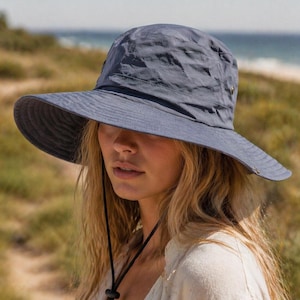 May include: A blue sun hat with a wide brim and a chin strap. The hat is made of a lightweight, textured fabric. The background shows a beach and the sea.