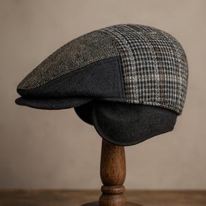 Op de afbeelding: Een tweed newsboy-pet met een donkergrijze oorbedekking. De pet heeft een ruitpatroon in grijstinten, bruin en zwart. De pet wordt getoond op een houten hoedenstandaard.