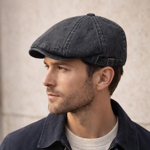May include: A dark gray denim flat cap with a small brim and adjustable strap. The cap is made of a textured fabric and has a button on top. The cap is worn by a person.