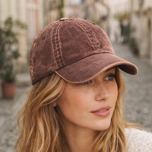 Puede incluir: Una gorra de béisbol de pana marrón con una construcción de seis paneles, visera curva y un botón en la parte superior. La tela presenta una textura acanalada vertical distintiva con un aspecto ligeramente descolorido y desgastado.