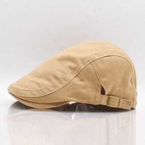 May include: A tan-colored, flat cap with a curved brim. The cap has a structured design with a stitched seam detail and an adjustable strap with a metal buckle at the back. The hat is made of a durable fabric.
