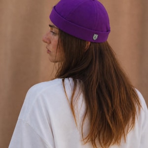 Könnte beinhalten: Eine leuchtend violette Beanie-Mütze mit einem kleinen Emblem an der Seite. Die Mütze wird von einer Person getragen, die ein weißes T-Shirt trägt. Der Hintergrund ist einfarbig warmbraun.