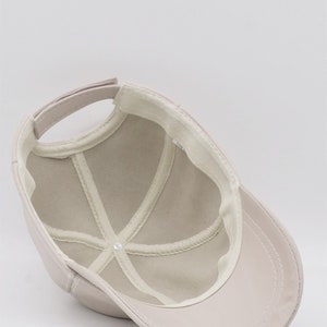 May include: A light beige baseball cap, viewed from the inside, showing the crown's structure and the adjustable strap. The cap features a curved brim and a white interior lining with stitched details. The cap is made of a smooth, solid-colored material.