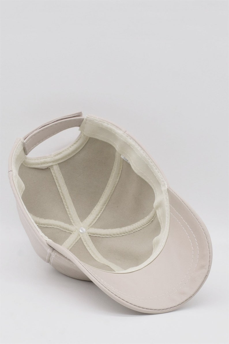 May include: A light beige baseball cap, viewed from the inside, showing the crown's structure and the adjustable strap. The cap features a curved brim and a white interior lining with stitched details. The cap is made of a smooth, solid-colored material.