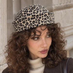 May include: A brimless beanie hat featuring a tan and black leopard print pattern. The hat is constructed with a thick, folded cuff around the base and has a rounded crown. The fabric appears to have a textured, corduroy-like finish throughout.