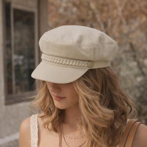 Op de afbeelding: Een beige newsboy-pet met een gevlochten band en een klein knoopdetail. De pet heeft een korte klep en een gestructureerde kroon. De hoed is gemaakt van een zachte, getextureerde stof. De pet is een stijlvol accessoire.