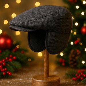 Puede incluir: Gorra de estilo newsboy gris oscuro con estampado de espiga y orejeras, exhibida en un soporte de madera. La gorra tiene una corona redondeada y una visera rígida. Las orejeras ofrecen calidez. Decoraciones navideñas en el fondo.