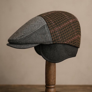 Könnte beinhalten: Eine Tweed-Schiebermütze mit Ohrenklappen, ausgestellt auf einem Holzständer. Die Mütze hat ein Patchwork-Design mit grauen, braunen und schwarzen Abschnitten. Der braune Abschnitt hat ein Karomuster mit rosa Akzenten.