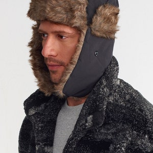 May include: A black winter hat with ear flaps and a faux fur trim. The hat is worn by a person wearing a grey shirt and a black and grey patterned jacket. The hat has a button closure on the ear flaps.