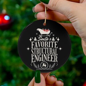 May include: A black ceramic Christmas ornament with gold string, held by a person. The ornament features white text that reads "Santa's Favorite Structural Engineer" with a sleigh and reindeer graphic. Red Christmas ornaments and a green tree are in the background.