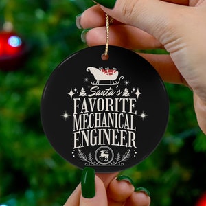 Op de afbeelding: Een zwarte keramische kerstversiering met een gouden draad, met de tekst "Santa's Favorite Mechanical Engineer" in het wit. Het ontwerp omvat een slee, sterren en een rendierembleem. De versiering wordt voor een kerstboom gehouden.