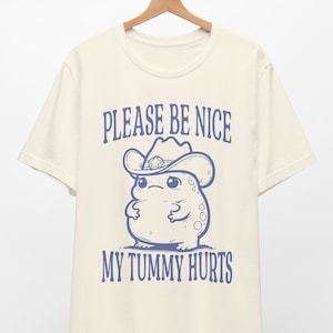 Op de afbeelding: Een crèmekleurig T-shirt met een cartoon kikker die een cowboyhoed draagt. De tekst "Please be nice, my tummy hurts" is in blauw op het shirt gedrukt.