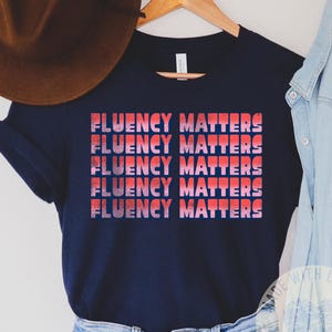 Könnte beinhalten: Ein marineblaues T-Shirt mit dem Text "FLUENCY MATTERS" in einem gestapelten Design im Retro-Stil. Der Text ist in Rot und Rosa gehalten. Das Shirt wird mit Jeansshorts und einem braunen Hut kombiniert.
