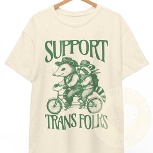以下が含まれることがあります： ベージュのTシャツに、緑色の動物2匹が自転車に乗っているグラフィックがプリントされています。グラフィックの下には、緑色の文字で「SUPPORT TRANS FOLKS」と書いてあります。