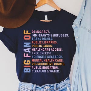 Könnte beinhalten: Ein dunkelblaues T-Shirt mit dem Text BIG FAN OF, gefolgt von einer Liste sozialer Anliegen wie Demokratie, Einwanderer, Flüchtlinge, Trans-Rechte, Bibliotheken, öffentliches Land, Gesundheitsversorgung, freie Meinungsäußerung und saubere Luft.