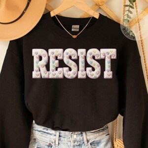 May include: Black sweatshirt with the word "RESIST" in large, puffy letters. The letters are filled with pastel-colored hearts. The sweatshirt is paired with denim shorts and a gold necklace.