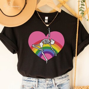 Può includere: Una t-shirt nera a maniche corte con la grafica di uno scheletro bianco che danza all'interno di un grande cuore rosa sovrapposto a un arcobaleno colorato. La maglietta è abbinata a pantaloncini di jeans azzurri con dettagli strappati.