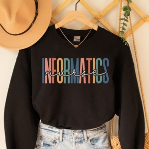 Può includere: Felpa nera con il testo "INFORMATICS nurse" in colori arcobaleno. La felpa è su un appendiabiti ed è abbinata a pantaloncini di jeans e una borsa con frange.