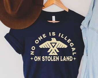 Może przedstawiać: Koszulka w kolorze granatowym z żółtym nadrukiem stylizowanego orła i tekstem "No one is illegal on stolen land".
