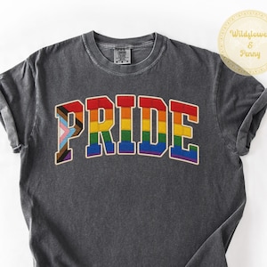 Puede incluir: Una camiseta gris oscuro con la palabra "PRIDE" en colores del arcoíris en la parte delantera. La palabra está en un estilo de letras en negrita, con un contorno blanco. La camiseta tiene mangas cortas y cuello redondo.