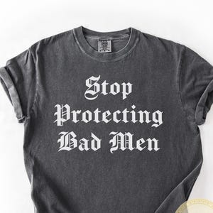 May include: A dark gray t-shirt with the text "Stop Protecting Bad Men" in a gothic font. The shirt has rolled-up sleeves and a round neckline. A small circular logo and a tag with the word "Pepper" are visible.