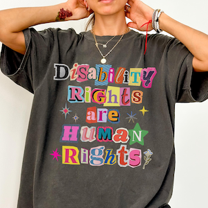 May include: A charcoal gray short-sleeve t-shirt featuring the multi-colored text "Disability Rights are Human Rights" in a collage-style font. The design includes small star graphics and a floral icon, printed on a relaxed-fit cotton garment.