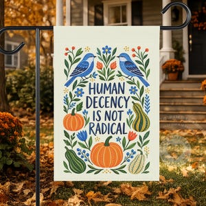May include: A rectangular garden flag featuring a cream background with the text Human Decency Is Not Radical in dark blue. The design includes two blue birds, orange pumpkins, green gourds, and colorful floral accents arranged in a symmetrical pattern.