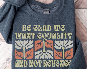 以下が含まれることがあります： レトロなフォントで「Be glad we want equality and not revenge」と書かれた濃いグレーのスウェットシャツ。テキストはオレンジと白の花のパターンで囲まれています。