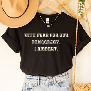 Può includere: Maglietta nera con testo bianco che recita "Con paura per la nostra democrazia, dissento."