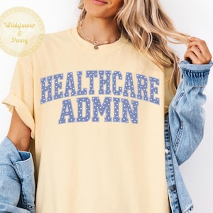 以下が含まれることがあります： ペールイエローの半袖Tシャツで、青いカレッジフォントに白い水玉模様の「HEALTHCARE ADMIN」という文字がプリントされています。Tシャツは青いデニムジャケットと一緒に着用されています。左上の円形ロゴには「Wildflower & Penny」と書かれています。