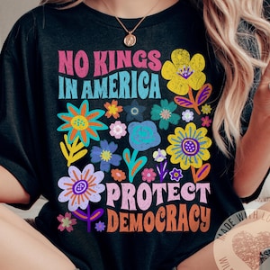Puede incluir: Camiseta negra con un dise&ntilde;o floral colorido y el texto "No Kings in America Protect Democracy". La camiseta tambi&eacute;n tiene un peque&ntilde;o logotipo que dice "Made with Love by Wildflower & Penny".