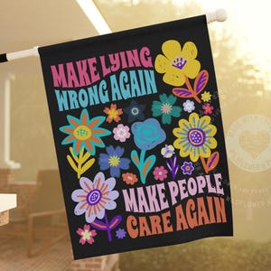 Może przedstawiać: Czarna flaga z napisem "MAKE LYING WRONG AGAIN MAKE PEOPLE CARE AGAIN" w kolorowych literach w stylu retro. Flaga jest ozdobiona różnymi kolorowymi ilustracjami kwiatów. Flaga wisi na zewnątrz na białym słupie.