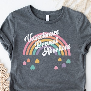 Pu&ograve; includere: Una maglietta grigio scuro con una grafica arcobaleno e il testo "Vasectomies Prevent Abortions". L'arcobaleno &egrave; composto da diversi colori, tra cui arancione, giallo, verde, blu, viola e rosa. Ci sono piccoli cuori di vari colori che pendono dall'arcobaleno.