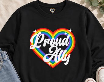 Può includere: Felpa nera con un design a cuore arcobaleno e la scritta "Proud Ally" in bianco. La felpa è abbinata a jeans blu e scarpe da ginnastica bianche. Il design presenta un arcobaleno a forma di cuore con stelle bianche.